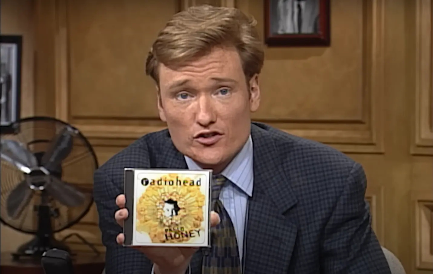 elevision host in a checkered suit holding up Radiohead’s 'Pablo Honey' CD, featuring the album’s distinctive yellow floral cover art, during a talk show segment with vintage studio background and fan visible.